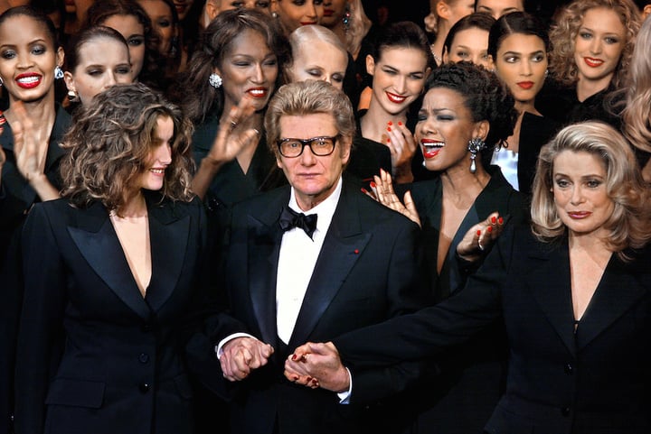 PARIS, FRANCE:  French designer Yves Saint-Laurent (C) salutes the crowd with French model Laetitia Casta (L) and French actress Catherine Deneuve (R) at the Centre Georges Pompidou art gallery in Paris, where the last Haute-Couture show ever of the designer took place. Hours before unveiling his last collection, innovative designer Yves Saint-Laurent said his love affair with women was not over and hinted he had other cards up his sleeve. (Photo credit should read JEAN-PIERRE MULLER/AFP/Getty Images)